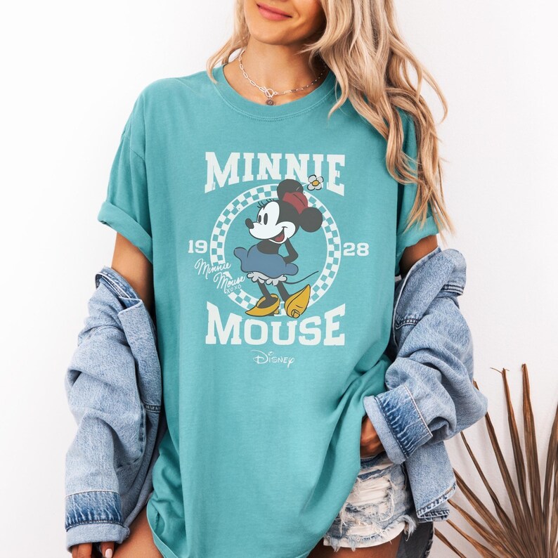 Puede incluir: Una camiseta de manga corta color verde azulado con un gr&aacute;fico de Minnie Mouse dentro de un c&iacute;rculo a cuadros. El texto dice MINNIE MOUSE 1928 con un peque&ntilde;o logotipo de Disney en la parte inferior. La camiseta tiene cuello redondo y corte holgado.