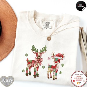 Puede incluir: Camiseta de color marfil con dos diseños de renos con patrones de patchwork en rojo, verde y beige. Los renos tienen cuernos verdes y están decorados con copos de nieve. La camiseta tiene cuello redondo y mangas cortas.