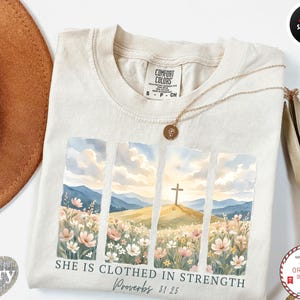 Puede incluir: Camiseta de color marfil con un diseño de paisaje en acuarela que presenta una cruz en una colina, flores y el texto "SHE IS CLOTHED IN STRENGTH Proverbs 31:25". También se ven un collar dorado y un sombrero marrón.