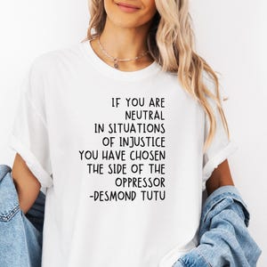 May include: White t-shirt featuring the quote "IF YOU ARE NEUTRAL IN SITUATIONS OF INJUSTICE YOU HAVE CHOSEN THE SIDE OF THE OPPRESSOR -DESMOND TUTU" in black. Worn with a denim jacket.