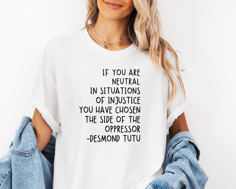 May include: White t-shirt featuring the quote "IF YOU ARE NEUTRAL IN SITUATIONS OF INJUSTICE YOU HAVE CHOSEN THE SIDE OF THE OPPRESSOR -DESMOND TUTU" in black. Worn with a denim jacket.