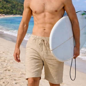 May include: A person on a sandy beach wearing beige shorts and holding a white surfboard with green and blue fins. The background shows a turquoise sea and a green hillside.
