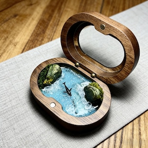 May include: An open, oval-shaped wooden box. Inside, a miniature ocean scene depicts a black orca in blue water, with green moss-covered rocks. The box is open, showcasing the detailed interior design.