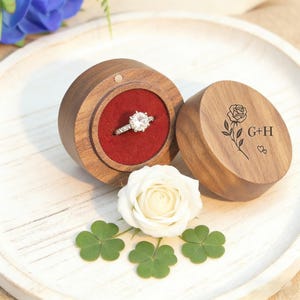 Op de afbeelding: Een ronde houten ringdoos met een rode fluwelen binnenkant bevat een diamanten verlovingsring. De doos is open, met het deksel ernaast. Het deksel is gegraveerd met een roos en de initialen "G+H". Een witte roos en klavers zijn in de buurt geplaatst.
