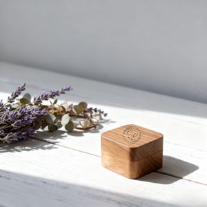 Op de afbeelding: Een kleine, vierkante houten doos met een scharnierend deksel. De bovenkant van de doos is gegraveerd met initialen en een datum. Links van de doos ligt een boeket lavendel en groen. De doos staat op een wit oppervlak.