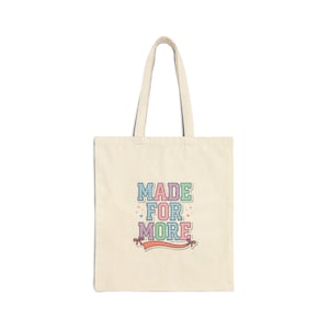 May include: A beige canvas tote bag featuring the text "MADE FOR MORE" in a multi-colored, checkered block font. The design includes small heart and star accents, with a coral-colored ribbon graphic underneath the text on the front panel.