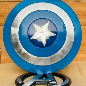 May include: A Captain America shield replica, featuring a blue, silver, and white color scheme. The shield has a central white star on a blue background, surrounded by silver and blue concentric circles. An Avengers logo is at the base.
