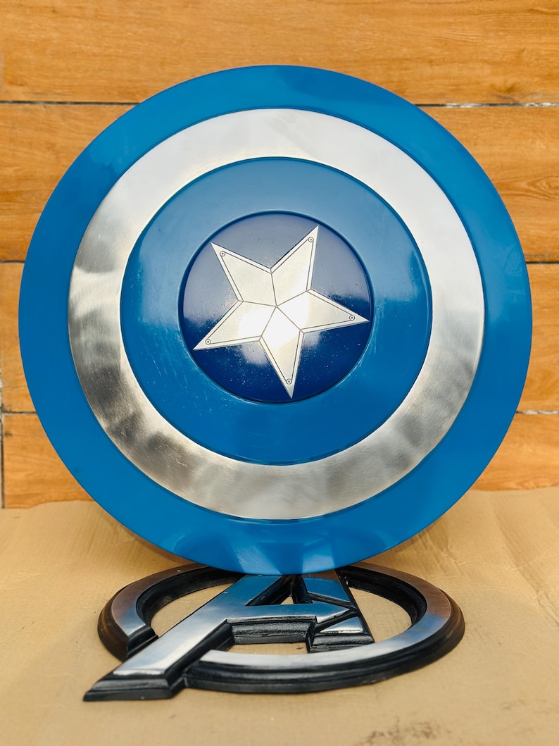 May include: A Captain America shield replica, featuring a blue, silver, and white color scheme. The shield has a central white star on a blue background, surrounded by silver and blue concentric circles. An Avengers logo is at the base.
