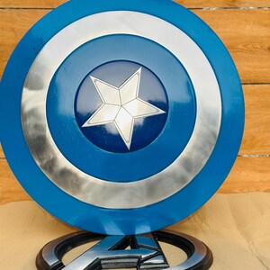 May include: A blue, silver, and white Captain America shield with a central white star design. The shield is round with a blue outer ring, a silver middle ring, and a blue center. Below the shield is a silver and black Avengers logo.