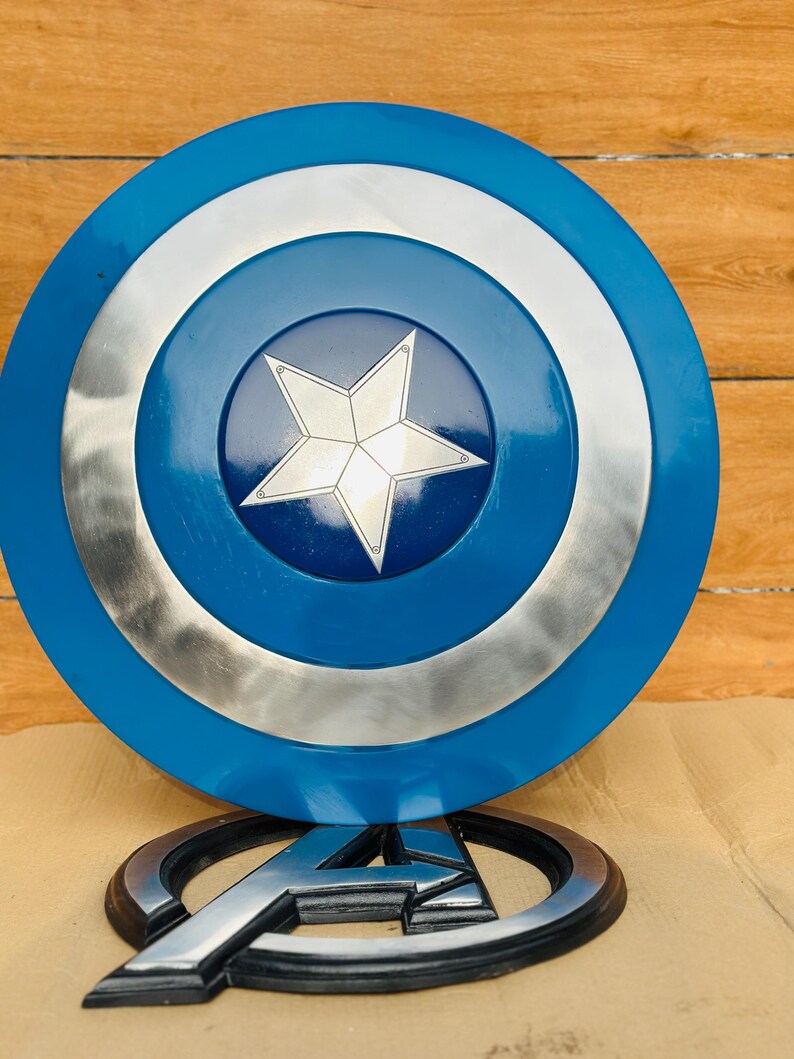 May include: A blue, silver, and white Captain America shield with a central white star design. The shield is round with a blue outer ring, a silver middle ring, and a blue center. Below the shield is a silver and black Avengers logo.