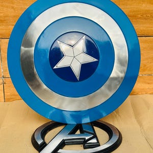 May include: A Captain America shield with a blue, silver, and white design. The shield has a central star emblem and is displayed with an Avengers logo.