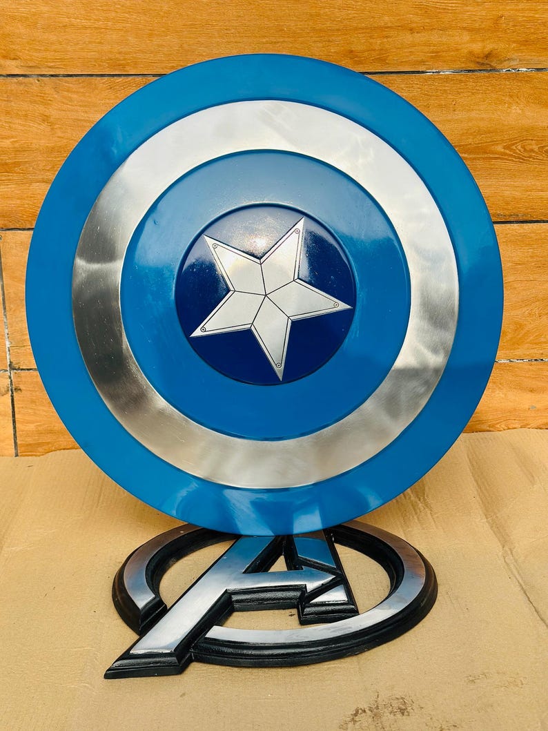 May include: A Captain America shield with a blue, silver, and white design. The shield has a central star emblem and is displayed with an Avengers logo.