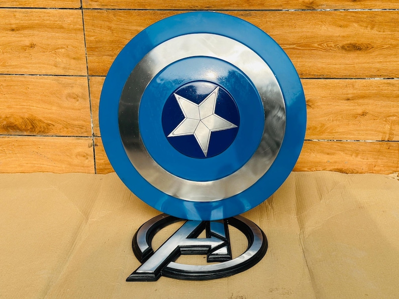 May include: A blue and silver Captain America shield with a white star in the center. The shield is round with concentric circles. Below the shield is a silver and black Avengers logo.