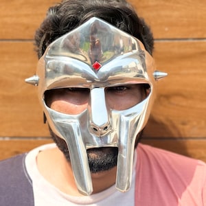 May include: A silver mask with a unique design, featuring a prominent nose and mouth section, and a red gem. The mask has a reflective surface and is worn by a person. The mask has a unique, futuristic aesthetic.