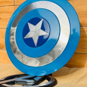 May include: A blue and silver Captain America shield with a white star in the center. The shield has concentric circles of blue and silver. The Avengers logo is in the foreground.