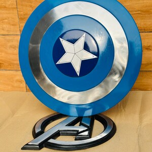 May include: A blue and silver Captain America shield with a white star in the center. Below the shield is a silver and black Avengers logo.