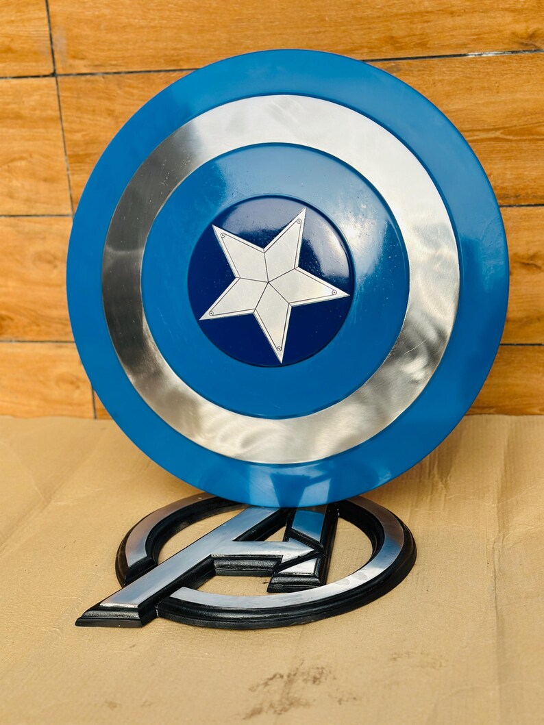 May include: A blue and silver Captain America shield with a white star in the center. Below the shield is a silver and black Avengers logo.