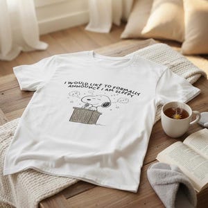 Könnte beinhalten: Weißes T-Shirt mit Snoopy-Grafik und dem Text "I would like to formally announce I am sleepy." Die Grafik zeigt Snoopy an einem Rednerpult. Eine Tasse Tee, ein Buch und Hausschuhe liegen auf einem Holztisch.