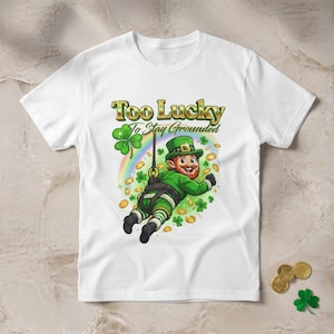May include: White t-shirt featuring a cartoon leprechaun in a green outfit, suspended from a rope, with the text "Too Lucky To Stay Grounded." The design includes a rainbow, shamrocks, and gold coins, perfect for St. Patrick's Day.