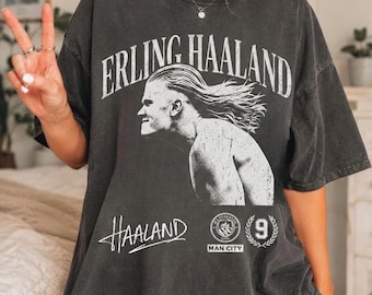 Op de afbeelding: Een antracietgrijs oversized T-shirt met een witte grafische print van Erling Haaland in profiel. De tekst luidt ERLING HAALAND, HAALAND, MAN CITY en het cijfer 9 in een cirkelvormig embleem. Het shirt heeft een ronde hals en korte mouwen.