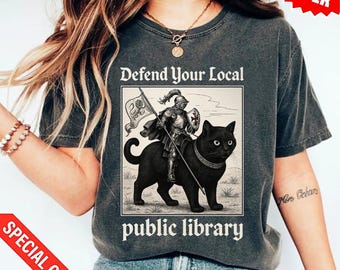 Op de afbeelding: Donkergrijs t-shirt met de tekst "Defend Your Local Public Library". De afbeelding toont een ridder op een zwarte kat met een vlag. Het t-shirt heeft een vintage, versleten uitstraling.