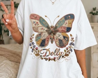 Op de afbeelding: Een wit t-shirt met korte mouwen en een grote afbeelding van een vlinder met vleugels in patchworkstijl in gedempte tinten blauw, roze en bruin. Onder de vlinder staat de naam Dolly in een cursief lettertype, omringd door bloemaccenten.