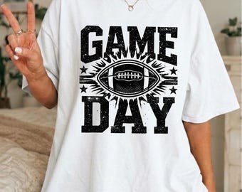 Op de afbeelding: Een wit T-shirt met korte mouwen en een zwarte grafische print met de tekst GAME DAY in een verweerd lettertype. In het midden staat een gedetailleerde illustratie van een American football, omringd door sterren en stralen.