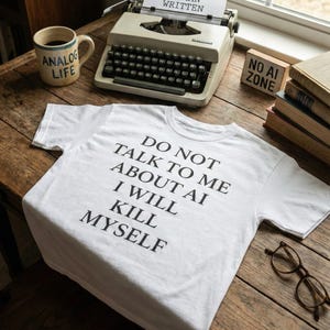 Könnte beinhalten: Weißes T-Shirt mit dem Text "DO NOT TALK TO ME ABOUT AI I WILL KILL MYSELF." Eine alte Schreibmaschine mit einem Papier mit der Aufschrift "HUMAN WRITTEN" steht auf einem Holztisch, zusammen mit einer Tasse mit der Aufschrift "ANALOG LIFE."
