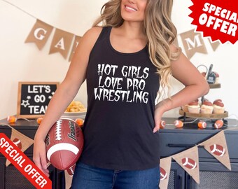 Op de afbeelding: Een zwart tanktop met de tekst "Hot Girls Love Pro Wrestling" in witte letters. Het tanktop wordt gedragen door een persoon die een voetbal vasthoudt.