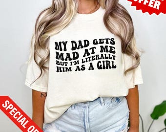 Op de afbeelding: Crèmekleurig T-shirt met de zwarte tekst "MY DAD GETS MAD AT ME BUT I'M LITERALLY HIM AS A GIRL". Het shirt wordt gecombineerd met een spijkerbroek. Een rode "SPECIAL OFFER" banner is zichtbaar.