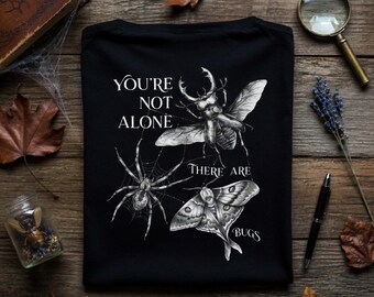 Op de afbeelding: Zwart t-shirt met een gedetailleerde illustratie van een spin, kever en mot. De tekst "YOU'RE NOT ALONE THERE ARE BUGS" is in het wit gedrukt. Het shirt wordt gepresenteerd op een houten oppervlak met decoratieve elementen.