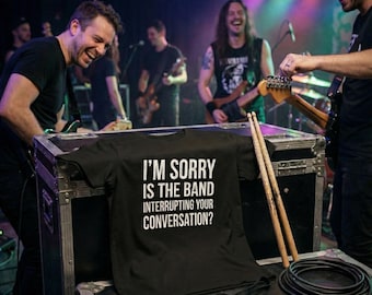 Op de afbeelding: Een zwart t-shirt met de tekst "I'M SORRY IS THE BAND INTERRUPTING YOUR CONVERSATION?" wordt op een podium getoond. Drumstokken en een gitaarkabel staan op de voorgrond. Op de achtergrond is een band te zien die op het podium optreedt.