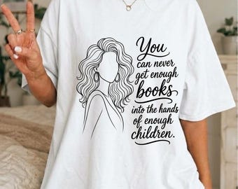 Op de afbeelding: Een wit T-shirt met korte mouwen en een zwarte lijntekening van een persoon met lang golvend haar en de tekst: You can never get enough books into the hands of enough children. Het shirt heeft een ronde hals en een losse pasvorm.