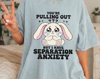 Op de afbeelding: Een lichtblauw T-shirt met korte mouwen en een afbeelding van een huilend wit konijn met roze oren. De tekst luidt: YOU'RE PULLING OUT ??? BUT I HAVE SEPARATION ANXIETY. Het shirt heeft een ronde hals en een losvallend, ontspannen model.
