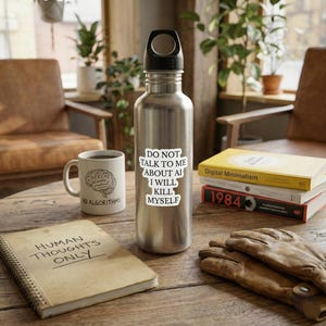 Könnte beinhalten: Eine silberne Wasserflasche mit schwarzem Deckel und einem Aufkleber mit der Aufschrift "DO NOT TALK TO ME ABOUT AI I WILL KILL MYSELF." Daneben befindet sich eine weiße Tasse mit einer Gehirngrafik und den Worten "NO ALGORITHMS." Ein Notizbuch mit "HUMAN THOUGHTS ONLY" ist ebenfalls zu sehen.