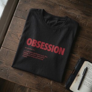 May include: A black crew neck t-shirt featuring the word OBSESSION in large red capital letters, followed by the phonetic spelling ob-ses-sion (n.) and a dictionary-style definition of the word printed in red text on the front of the garment.
