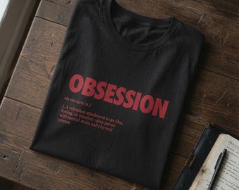Op de afbeelding: Een zwart t-shirt met ronde hals voorzien van het woord OBSESSION in grote rode hoofdletters, gevolgd door de fonetische spelling ob-ses-sion (n.) en een woordenboekachtige definitie van het woord in rode tekst op de voorzijde.