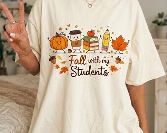 Op de afbeelding: Een crèmekleurig T-shirt met korte mouwen en een afbeelding van een lachende pompoen, koffiebeker, stapel boeken, potlood en esdoornblad die elkaars hand vasthouden. De tekst "Fall with my Students" staat eronder met verspreide herfstbladeren.
