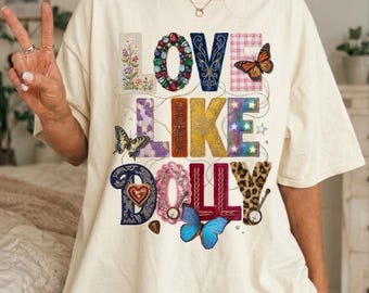 Op de afbeelding: Een off-white t-shirt met korte mouwen en de tekst LOVE LIKE DOLLY in kleurrijke, gedessineerde letters. Het ontwerp bevat bloemen-, edelsteen-, ruit-, luipaard- en stermotieven, geaccentueerd door vlinders.