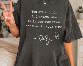 Op de afbeelding: Een antracietgrijs t-shirt met korte mouwen en witte tekst: You are enough. And anyone who tells you otherwise isn't worth your time - Dolly, vergezeld van een kleine witte lijntekening van een vlinder op de voorzijde.