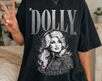 Op de afbeelding: Een zwart t-shirt met korte mouwen en het woord DOLLY in grote, sierlijke, zilverkleurige letters aan de bovenkant. Onder de tekst staat een gedetailleerde zwart-wit grafische illustratie van een vrouw met volumineus haar in een overhemd.