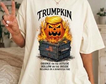 Op de afbeelding: Een off-white t-shirt met een afbeelding van een pompoen met blond haar in een brandende afvalcontainer. De tekst luidt bovenaan TRUMPKIN en onderaan ORANGE ON THE OUTSIDE HOLLOW ON THE INSIDE BELONGS IN A DUMPSTER FIRE.