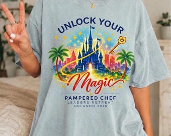 Op de afbeelding: Een lichtblauw t-shirt met een afbeelding van een blauw kasteel, palmbomen en een gouden sleutel. De tekst luidt UNLOCK YOUR Magic PAMPERED CHEF LEADERS RETREAT ORLANDO 2026. Het ontwerp bevat gele sterren en een kleurrijke skyline van een stad.