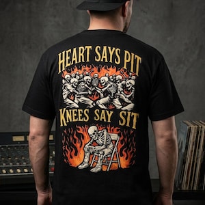 May include: A black short-sleeve t-shirt featuring a graphic print on the back. The design shows skeletons in a mosh pit above a skeleton sitting on a chair, surrounded by orange flames. Text reads: HEART SAYS PIT KNEES SAY SIT in yellow letters.