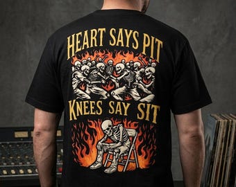 Op de afbeelding: Een zwart t-shirt met korte mouwen en een grafische print op de achterkant. Het ontwerp toont skeletten in een moshpit boven een skelet dat op een stoel zit, omringd door oranje vlammen. De tekst luidt: HEART SAYS PIT KNEES SAY SIT in geel.