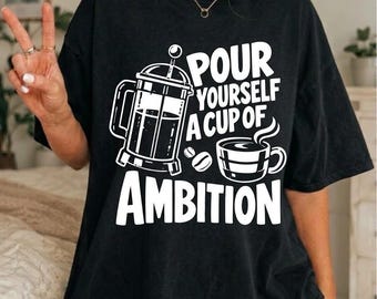 Op de afbeelding: Een zwart t-shirt met korte mouwen en een witte grafische print van een cafetière, een dampende kop koffie en twee koffiebonen. De tekst "POUR YOURSELF A CUP OF AMBITION" staat in een vetgedrukt, wit lettertype op de borst.