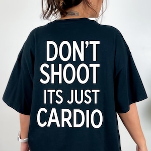 Może przedstawiać: Czarny t-shirt z krótkim rękawem z białym napisem na plecach o treści: DON'T SHOOT ITS JUST CARDIO. Koszulka ma okrągły dekolt oraz luźny, swobodny krój, co zapewnia prosty i nieskomplikowany wygląd do codziennego noszenia.