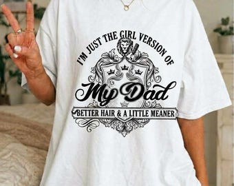 Op de afbeelding: Een wit T-shirt met korte mouwen en een zwarte grafische print van een leeuwenwapen met de tekst: I'M JUST THE GIRL VERSION OF My Dad BETTER HAIR & A LITTLE MEANER. Het ontwerp bevat sierlijke krullen en kleine kronen.