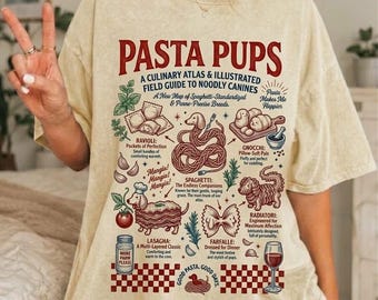 Op de afbeelding: Een beige t-shirt met korte mouwen en een grafische print met de titel PASTA PUPS. Het ontwerp bevat illustraties van verschillende pastavormen gecombineerd met honden, vergezeld van tekstlabels zoals Ravioli, Gnocchi, Spaghetti, Lasagna en Farfalle.