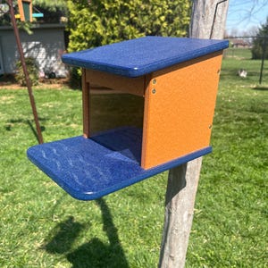 May include: A blue and orange bird feeder mounted on a tree. The feeder has a blue roof and a blue perch with a wave pattern. The sides are orange, and the feeder is designed to attract birds to a garden.